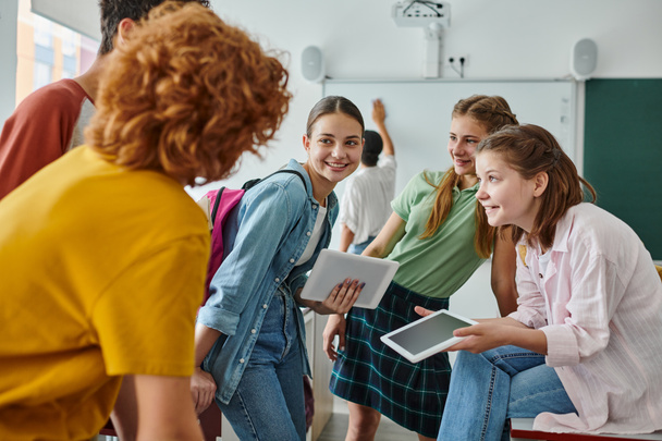 Sonrientes colegialas adolescentes sosteniendo tabletas digitales cerca de amigos borrosos en el aula en la escuela - Foto, Imagen
