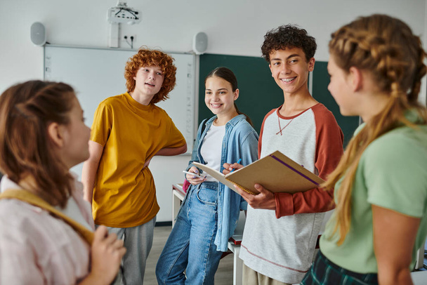 Escolar adolescente sonriente con cuaderno hablando con amigos mientras está de pie en el aula en la escuela - Foto, Imagen