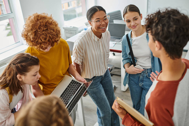 Рыжий мальчик показывает ноутбук однокласснику, африканский учитель Америки улыбается рядом со студентами - Фото, изображение