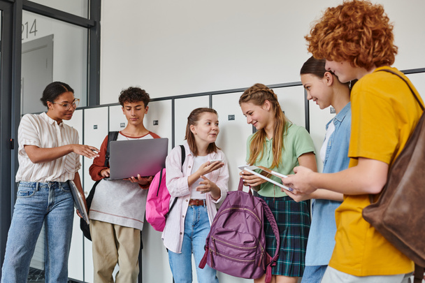 счастливые студенты-подростки, держащие устройства в школьном коридоре, африканская американка, стоящая рядом с парнем - Фото, изображение