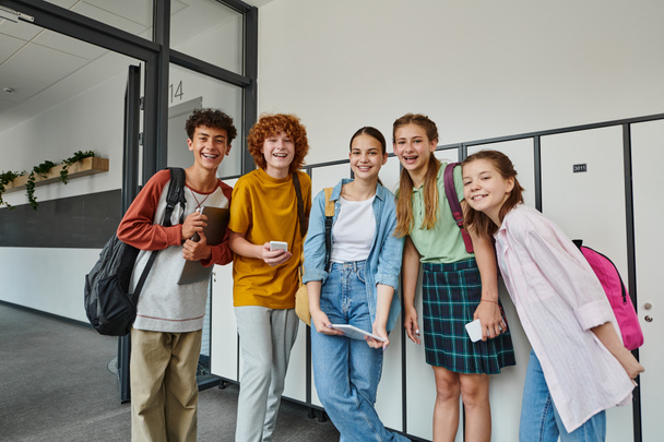 alegre adolescente estudiantes sosteniendo dispositivos y mirando a la cámara en el pasillo de la escuela, amigos adolescentes - Foto, Imagen