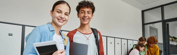 banner, tini osztálytársak kezében eszközök és nézi kamera az iskola folyosóján, boldog diákok - Fotó, kép