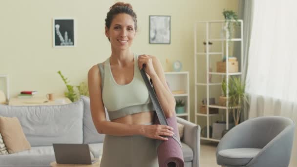 Ritratto medio di felice giovane donna caucasica in activewear stretto in posa per la fotocamera con stuoia di yoga rotolato sulla spalla a casa - Filmati, video