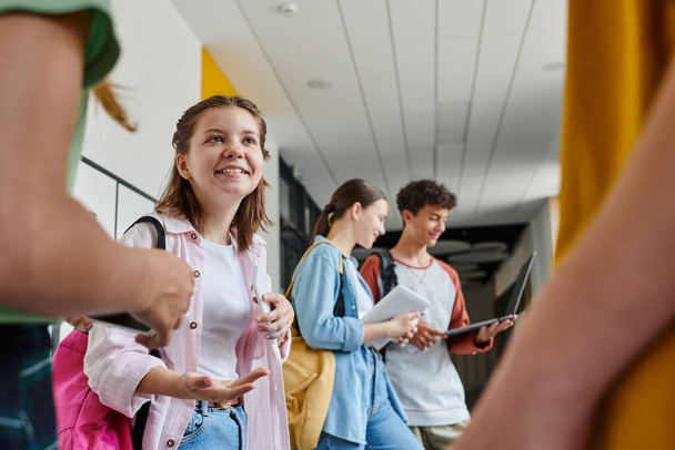 Вернуться в школу, счастливая девочка-подросток разговаривает с одноклассниками в коридоре, размытые студенты с помощью устройств - Фото, изображение