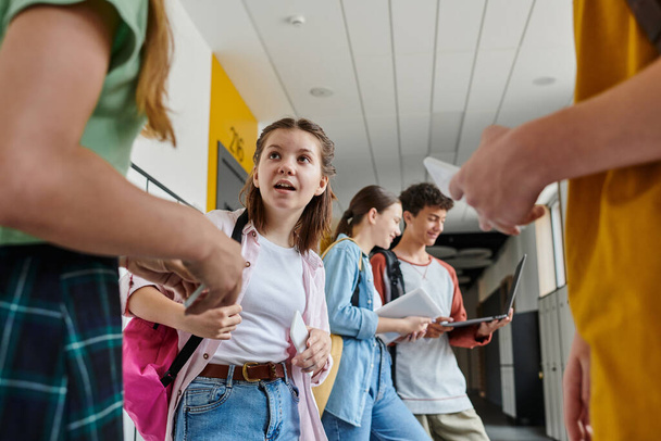 surprised schoolgirl looking at classmates in school hallway, teenage students with devices - Photo, Image