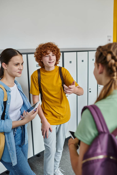 boldog fiú gesztikulál és chatel osztálytársával az iskola folyosóján, tinédzser diákok eszközök - Fotó, kép
