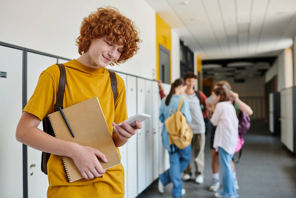 boldog vörös hajú iskolás fiú használ okostelefon és gazdaság notebook, elmosódott, diákok az iskola folyosóján - Fotó, kép