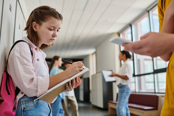 Сфокусированная школьница делает заметки, держит блокнот рядом с мальчиком, используя смартфон в школьном коридоре - Фото, изображение