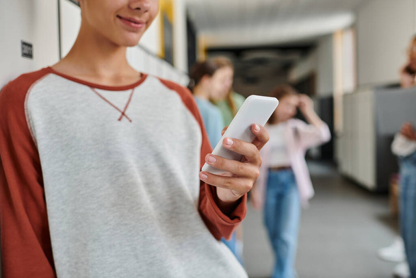 abgeschnittene Ansicht eines glücklichen Schuljungen mit Smartphone, SMS in der Pause auf dem Schulflur, Unschärfe - Foto, Bild