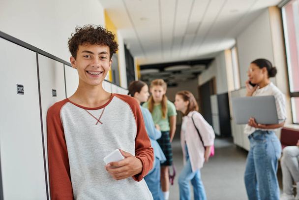 happy boy with braces holding smartphone and looking at camera during break in school hallway - Photo, Image