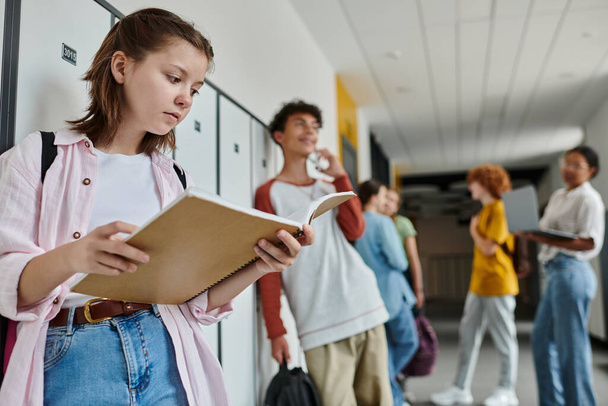 iskolás lány gazdaság notebook és áll közelében elmosódott és boldog tini osztálytárs, diákok, folyosón - Fotó, kép