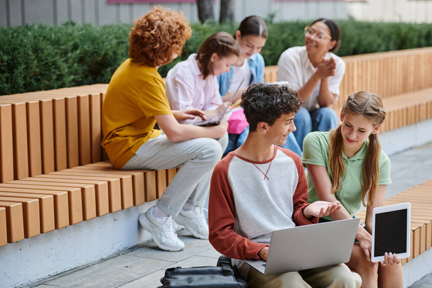 de vuelta a la escuela, niño adolescente feliz mostrando portátil a su compañero de clase, estudiantes y profesores al aire libre, diversidad - Foto, imagen