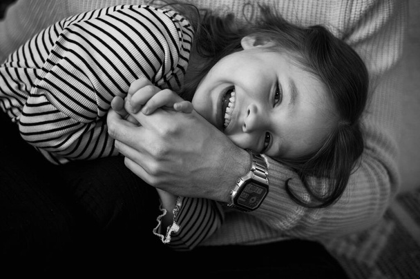 Boldog gyereknapot! Fekete-fehér fotó egy felismerhetetlen apáról és egy imádnivaló, nevető kislányról, akik együtt töltik a hétvégét otthon. Játékos gyermek lány és apa ölelés. - Fotó, kép