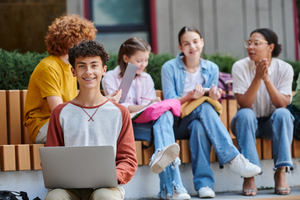 Снова в школу, счастливый мальчик-подросток смотрит в камеру, держа ноутбук, разнообразие, учителя и студенты - Фото, изображение