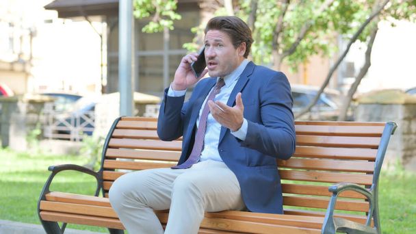 Empresario de mediana edad molesto discutiendo en el teléfono mientras está sentado al aire libre en un banco - Foto, imagen