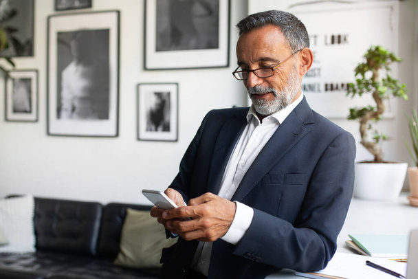 Pozitív jóképű kaukázusi vezető üzletember öltönyben és szemüvegben okostelefonon gépel a modern irodai térben. Alkalmazás távoli munkavégzésre, üzleti és pénzügyi ellenőrzés, startup - Fotó, kép