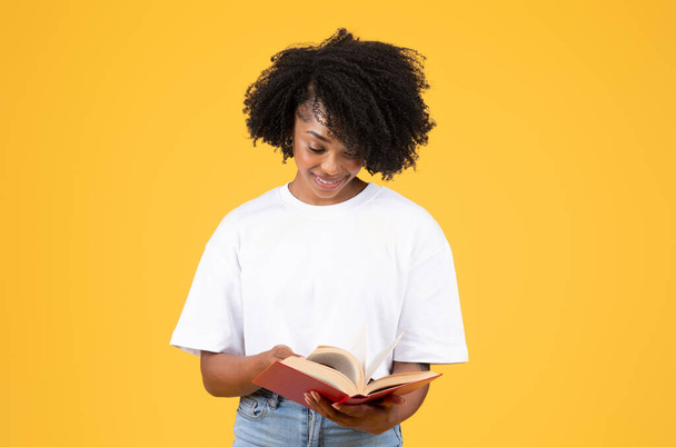 Positive young black curly lady in white t-shirt read book, enjoy study, isolated on orange background, studio. Ad and offer, education, knowledge and hobbies in free time - Photo, Image