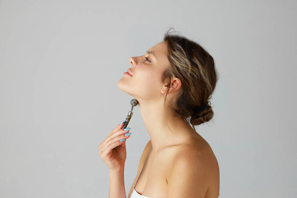 Beautiful young woman taking doing massage of neck with roller against grey studio background. Rejuvenating treatment. Concept of natural beauty, cosmetology and dermatology, skincare, cosmetics, ad - Fotó, kép