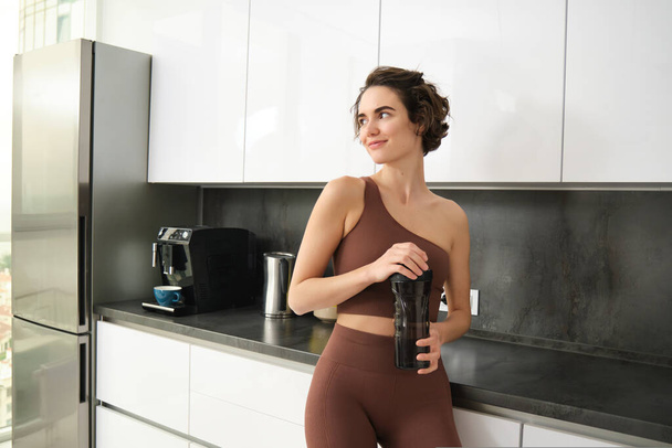 Sport et mode de vie sain. Portrait de femme de fitness souriante en vêtements de sport, debout près du comptoir de la cuisine à la maison, buvant dans une bouteille d'eau, se préparant à l'entraînement gymnase. - Photo, image