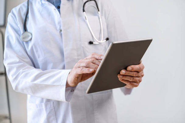 Widok dojrzałego lekarza trzymającego nowoczesną cyfrową tabletkę i korzystającego z aplikacji opieki zdrowotnej w szpitalu do zdalnej komunikacji z pacjentem. Recepta na leki, koncepcja konsultacji online - Zdjęcie, obraz