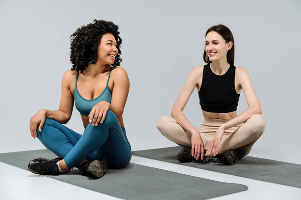 Zwei glückliche Freundinnen schauen sich im Fitnesscenter an, sitzen auf dem Boden auf der Yogamatte, lächeln breit und ruhen sich nach dem Training aus. Frauen machen Pilates isoliert auf grauem Hintergrund - Foto, Bild