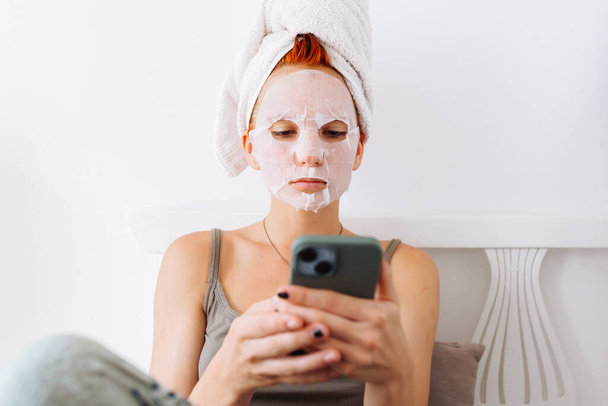 Retrato joven mujer pelirroja, adolescente, en máscara de sábana cosmética en la cara, con toalla en la cabeza, sobre fondo claro, en casa, en el salón de belleza, utilizando el teléfono móvil - Foto, Imagen