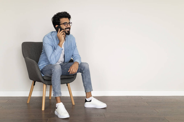 Smiling Young Indian Male Talking On Cellphone While Sitting In Armchair Indoors, Handsome Eastern Guy In Eyeglasses Enjoying Pleasant Phone Conversation, Talking With Friends Or Family, Copy Space - Foto, imagen