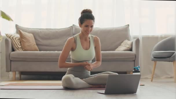 Young female yoga coach giving online video lesson via laptop, sitting on floor at home in lotus pose - Footage, Video