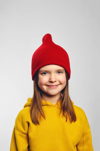 Vertical close up portrait of adorable hipster kid with pleasant smile, wearing yellow hoodie and red hat, standing isolated over white background - Zdjęcie, obraz