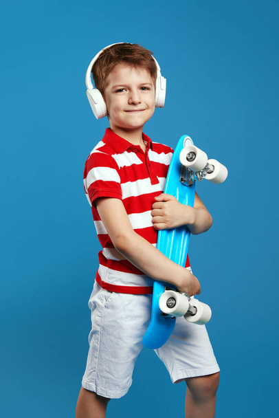 Niño alegre con traje de moda sosteniendo monopatín mientras escucha música a través de auriculares aislados sobre fondo de color azul. Retrato de estudio de niños. Concepto de estilo de vida infantil - Foto, Imagen