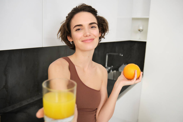Sağlıklı ve güzel genç bir kadın, size taze sıkılmış meyve suyu veriyor, portakal tutuyor ve gülümsüyor, idmandan, antrenmandan ve spor aktivitelerinden sonra C vitamini içiyor.. - Fotoğraf, Görsel