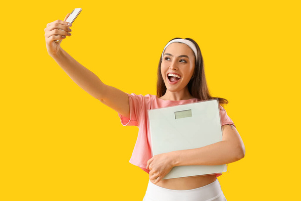 Mujer joven con básculas y teléfono móvil tomando selfie sobre fondo amarillo - Foto, imagen