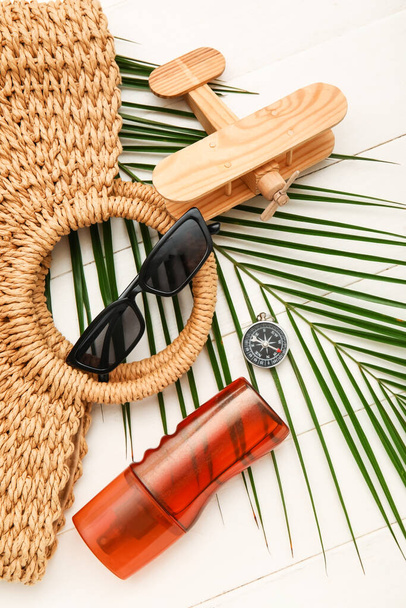 Güneş gözlüklü kompozisyon, hasır çanta, bir şişe güneş kremi ve beyaz ahşap arka planda palmiye yaprağı. - Fotoğraf, Görsel