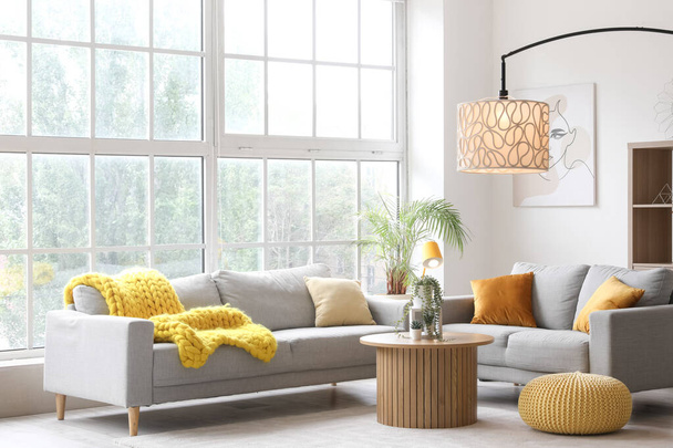 Interno di soggiorno luminoso con accoglienti divani grigi, tavolino e lampade luminose vicino a una grande finestra - Foto, immagini