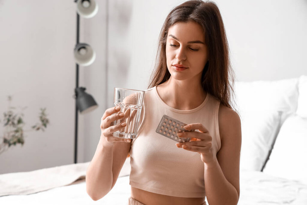 Fiatal nő egy pohár vízzel és fogamzásgátló tablettával a hálószobában - Fotó, kép