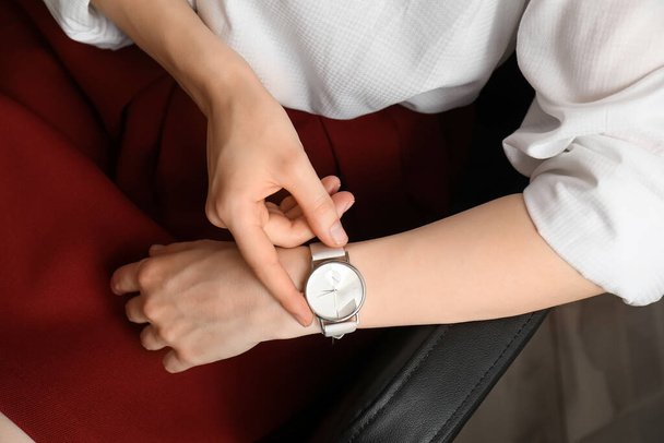 Junge Frau sitzt auf Sessel und schaut auf Armbanduhr im Zimmer, Nahaufnahme - Foto, Bild