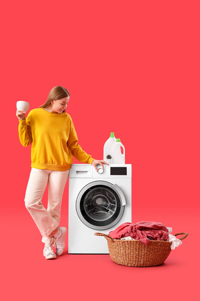 Pretty young woman with cup of tea setting up washing machine on red background - Fotó, kép