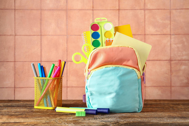 Цветной школьный рюкзак с ноутбуками, акварелями и чашкой маркеров на деревянном столе возле бежевой плитки - Фото, изображение