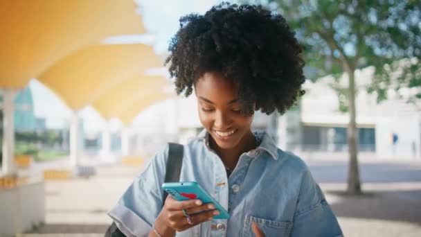 Chica alegre leyendo SMS mirando pantalla del teléfono inteligente en la calle soleada de cerca. Atractivo afroamericano adolescente mensajería por celular caminando campus. Feliz adolescente sonriente charlando en línea por móvil. - Metraje, vídeo
