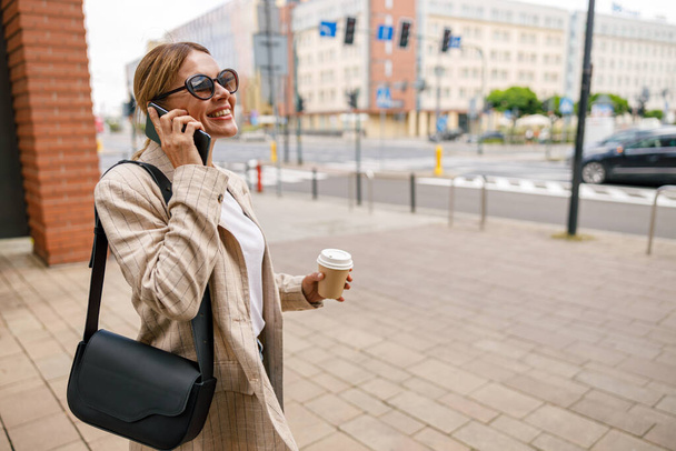 Smiling woman office worker is talking by phone while standing on modern building background  - Fotó, kép