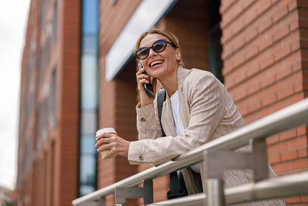 Smiling woman office worker is talking by phone while standing on modern building background  - Fotografie, Obrázek
