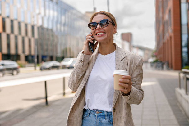 Smiling woman office worker is talking by phone while standing on modern building background  - Foto, imagen