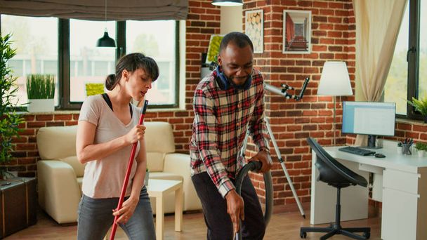Diverse life partners dancing and sweeping dust off floors, using vacuum cleaner and washing solution. Cheerful couple laughing and enjoying spring cleaning in apartment, household chores. - Photo, Image
