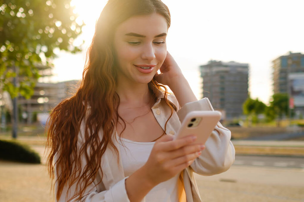 Romantikus csinos, fiatal, vörös hajú nő áll az utcán, és sms-ezik az okostelefonjával naplementekor - Fotó, kép