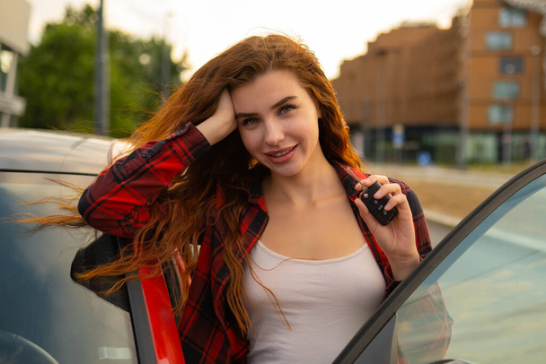 Com fechaduras vermelhas ardentes e um sorriso radiante, a jovem mulher vestindo camisa xadrez casual posa ao lado do carro vermelho e brinca com as chaves do carro em suas mãos. - Foto, Imagem