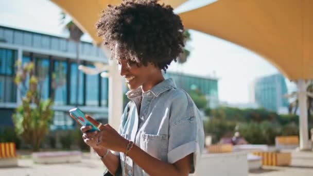Adolescent heureux regarder les médias sociaux sur smartphone marcher rue ensoleillée fermer. Joyeux afro-américaine fille souriant message de lecture sur téléphone portable à l'extérieur. mignon bouclé jeune femme textos sur aller. - Séquence, vidéo