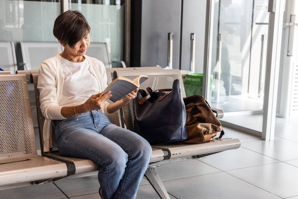 Edad media Mujer asiática con libro de lectura de equipaje para matar el tiempo mientras espera la salida en la terminal del aeropuerto - Foto, imagen