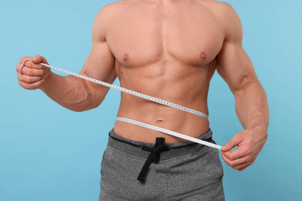 Athletic man measuring waist with tape on light blue background, closeup. Weight loss concept - 写真・画像