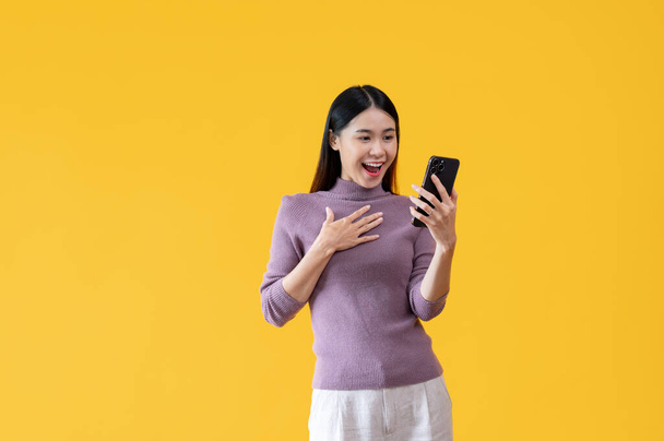 Une belle femme asiatique regarde son écran de smartphone avec un visage surprenant tout en se tenant sur un fond jaune isolé. choqué, étonné, wow, impressionné, satisfait - Photo, image