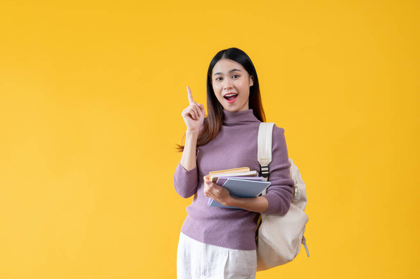 A beautiful and cheerful young Asian female college student with a backpack is carrying her books and pointing her finger up while standing against an isolated yellow background. - Foto, Bild